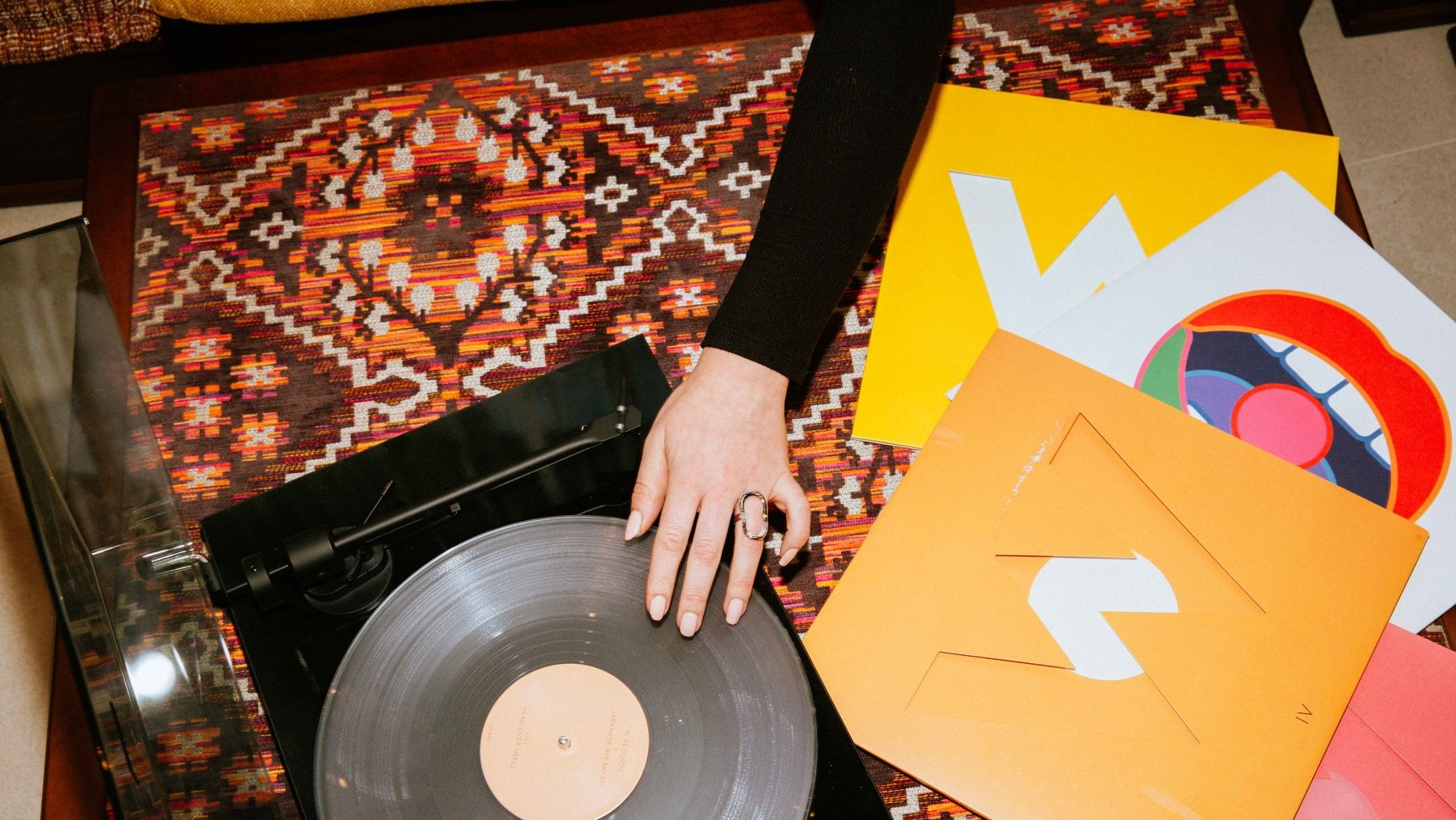 Hand playing a vinyl record beside colourful sleeves on a patterned rug