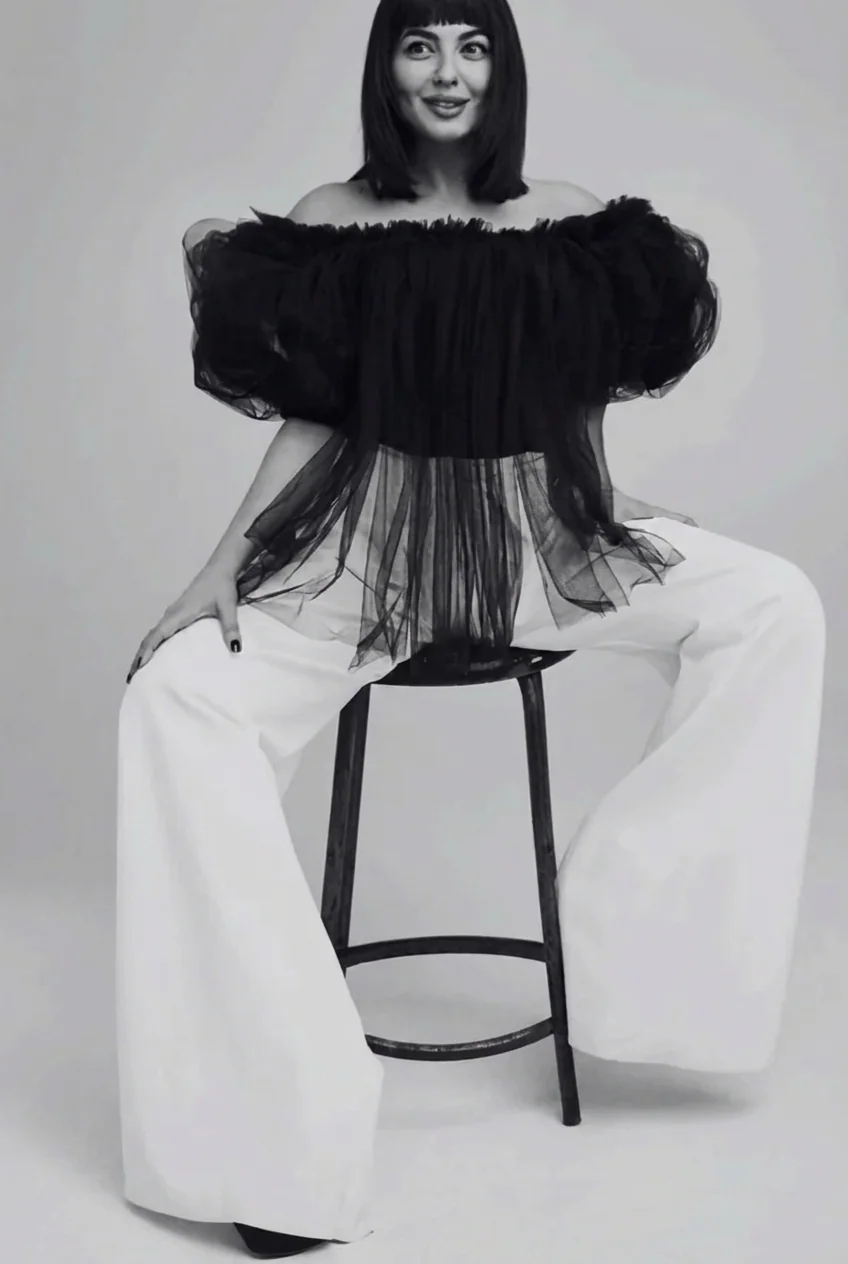 Portrait of Renata Kaveh seated on a stool in a black tulle top and white wide-leg trousers