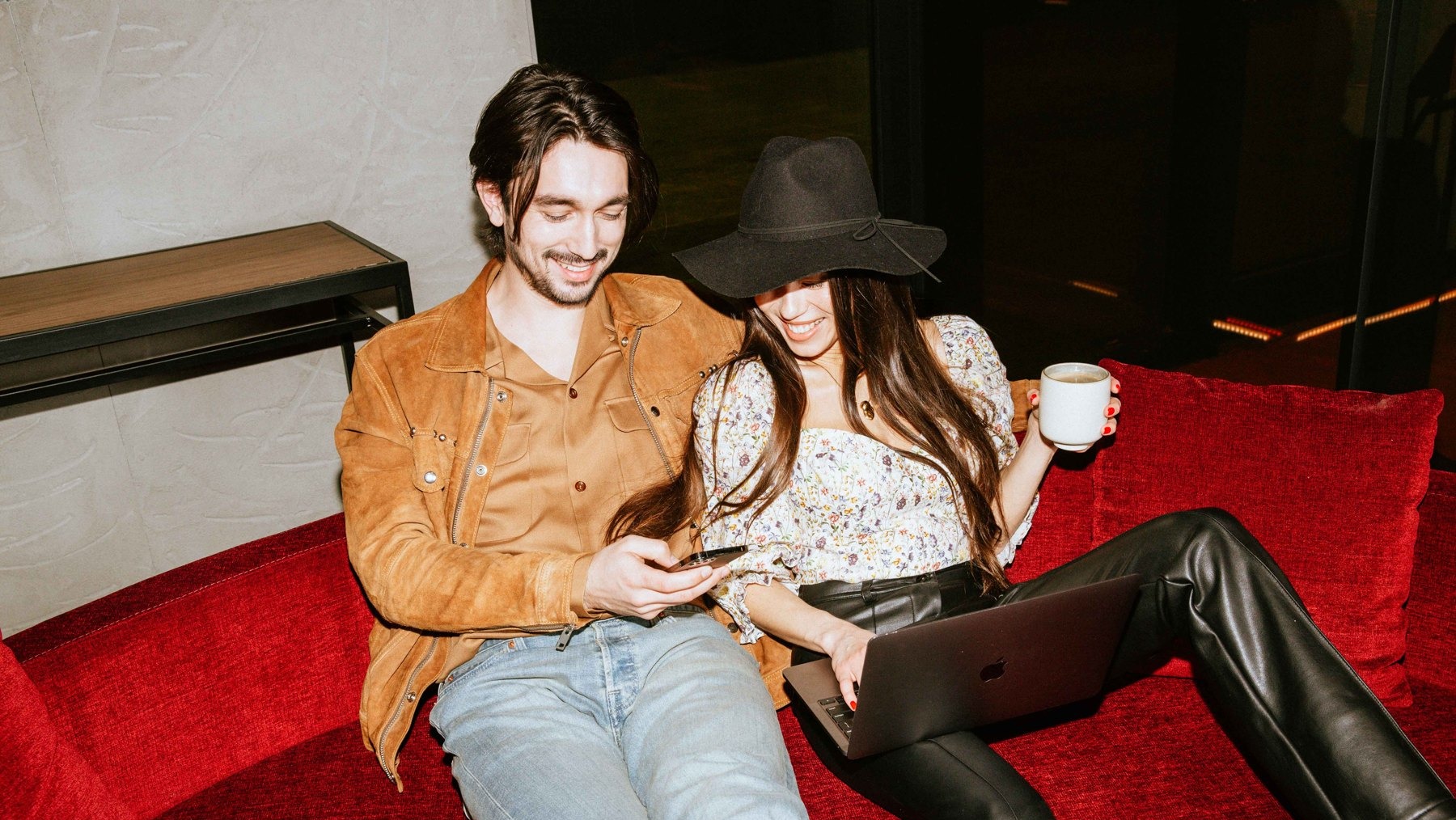 Couple laughing on a red velvet sofa with a laptop and coffee